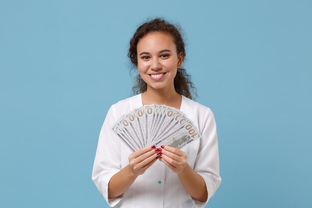 Sorridente medico afroamericano donna isolata su sfondo blu. Medico femminile in camice medico tenere fan di denaro contante in banconote in dollari. Concetto di medicina del personale sanitario. Simula lo spazio della copia.