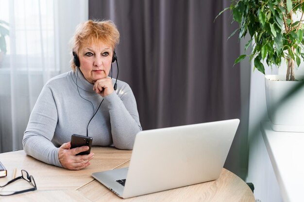 Sorridente moderna donna caucasica matura anni '60 in cuffia guarda il webinar online a casa. La femmina dai capelli grigi senior felice si diverte a studiare su internet. Concetto di tecnologia per anziani.