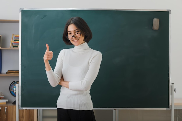 sorridente mostrando pollice in alto giovane insegnante femminile con gli occhiali in piedi davanti alla lavagna in classe