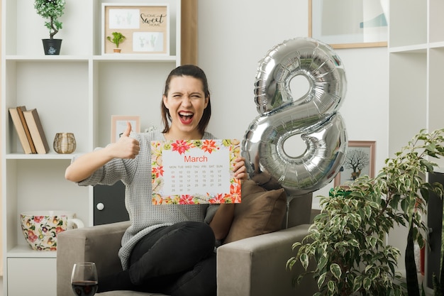 Sorridente mostrando pollice in su bella donna il giorno delle donne felici tenendo il calendario seduto sulla poltrona in soggiorno