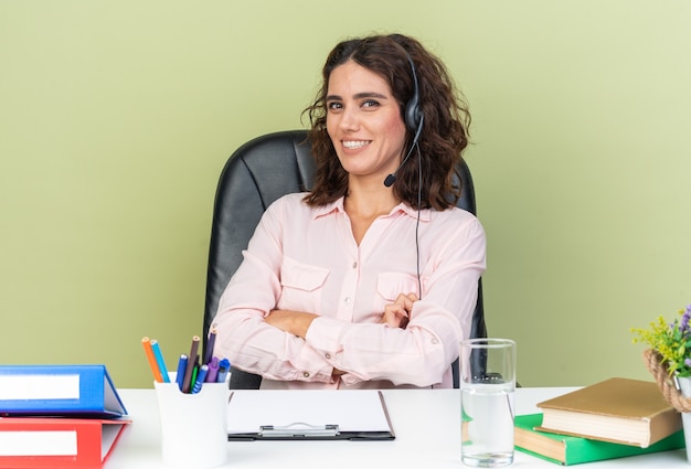 Sorridente operatore di call center femminile caucasica sulle cuffie seduto alla scrivania con strumenti da ufficio che incrociano le braccia isolate sul muro verde