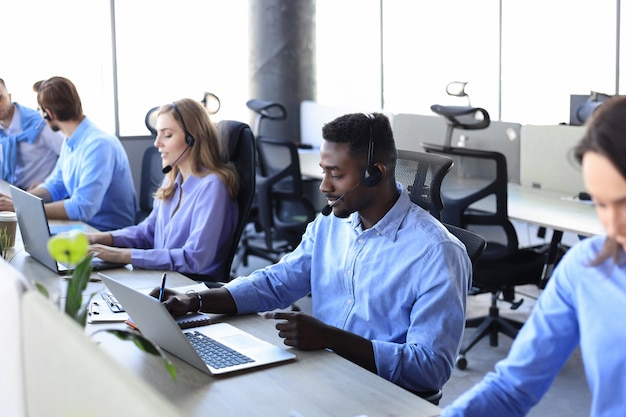 Sorridente operatore di call center maschio africano con cuffie seduto in un ufficio moderno con colleghi sullo sfondo, consulenza online.