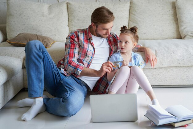 Sorridente padre e figlia seduti sul pavimento in soggiorno con laptop, lezioni di insegnamento.