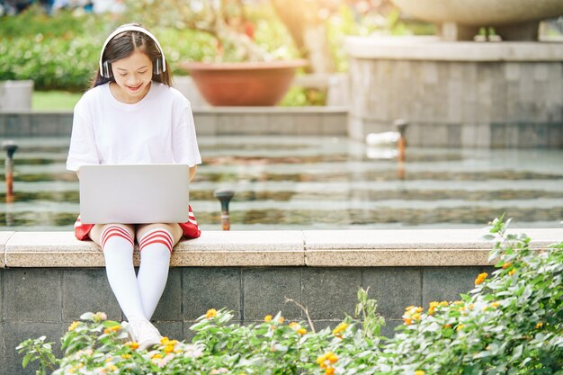 Sorridente ragazza adolescente asiatica seduta alla fontana e lavora al computer portatile, ascoltando musica e rispondendo alle e-mail