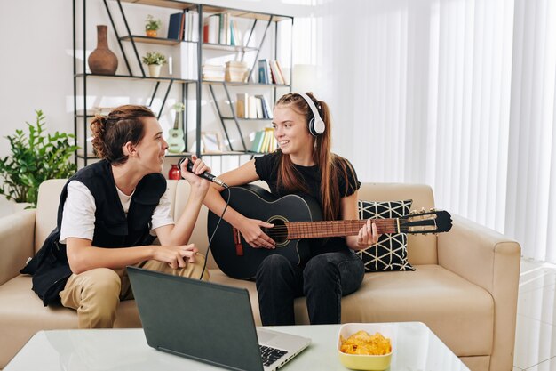 Sorridente ragazza adolescente in cuffie a suonare la chitarra quando la sua amica canta una canzone nel microfono