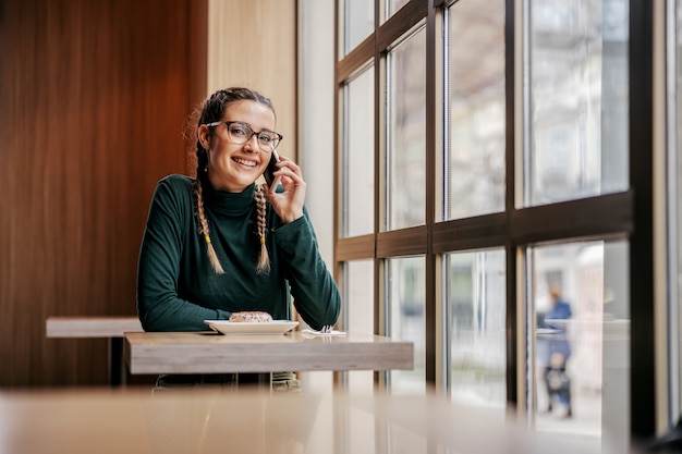 Sorridente ragazza adolescente seduto nella caffetteria accanto alla finestra e avere una conversazione telefonica. Concetto di telecomunicazione globale.