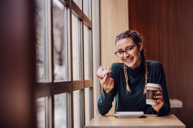 Sorridente ragazza adolescente seduto nella caffetteria, tenendo ciambella e tazza di caffè in pausa tra le classi.