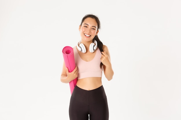 Sorridente ragazza asiatica felice di forma fisica in cuffie e abbigliamento sportivo, portare il tappetino in gomma per allenamento o yoga, ridendo spensierata su sfondo bianco.