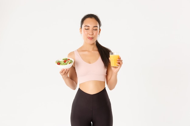 Sorridente ragazza asiatica fitness carino in activewear, leccandosi le labbra soddisfatte con una deliziosa insalata sana e succo d'arancia prima dell'allenamento in palestra.