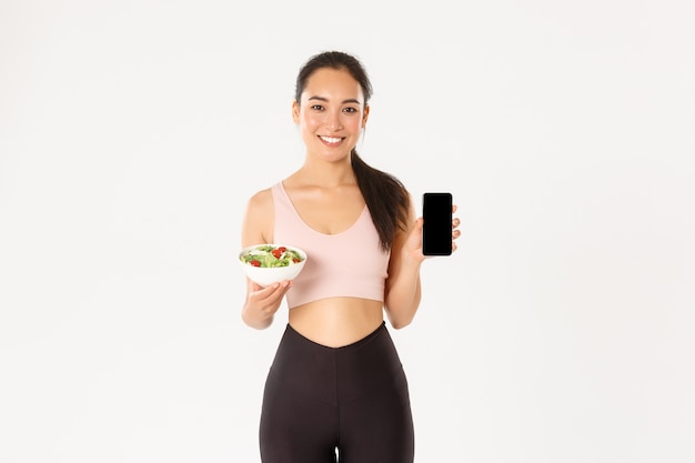 Sorridente ragazza asiatica snella e carina, allenatore di palestra che mostra insalata e schermo dello smartphone, consiglia di scaricare tracker dietetico o promemoria calorie.