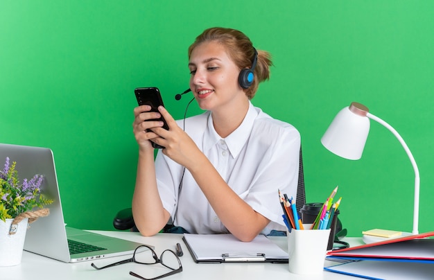 Sorridente ragazza bionda del call center che indossa l'auricolare seduto alla scrivania con strumenti di lavoro usando il suo telefono cellulare