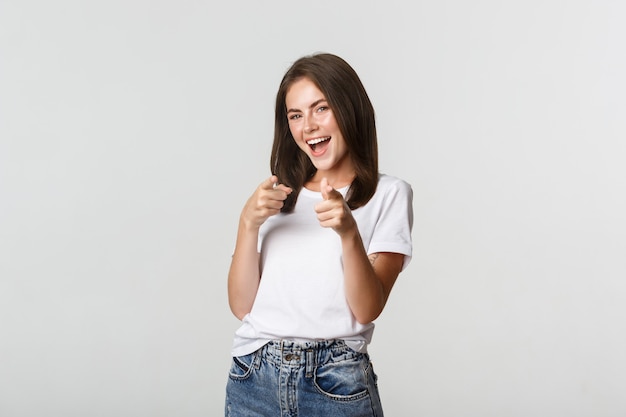 Sorridente ragazza bruna allegra che punta il dito alla macchina fotografica, congratularmi con te, lodando la buona scelta