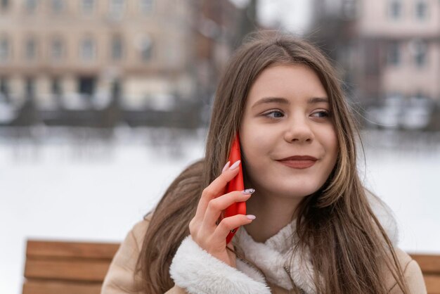 Sorridente ragazza carina si siede sulla panchina del parco e parla sullo smartphone Sorridente giovane donna in cappotto di pelle di pecora beige riposa all'esterno