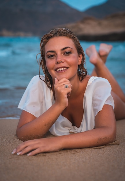 Sorridente ragazza caucasica bionda sdraiata sulla spiaggia in un costume da bagno bianco