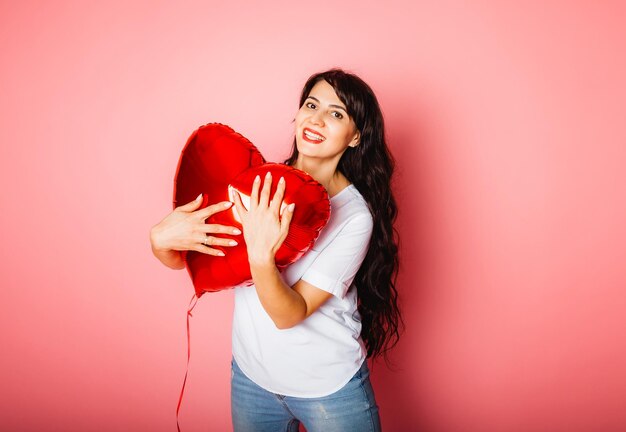 Sorridente ragazza caucasica soddisfatta in posa con un palloncino rosso a forma di cuore su sfondo rosa. San Valentino