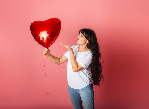 Sorridente ragazza caucasica soddisfatta in posa con un palloncino rosso a forma di cuore su sfondo rosa. San Valentino