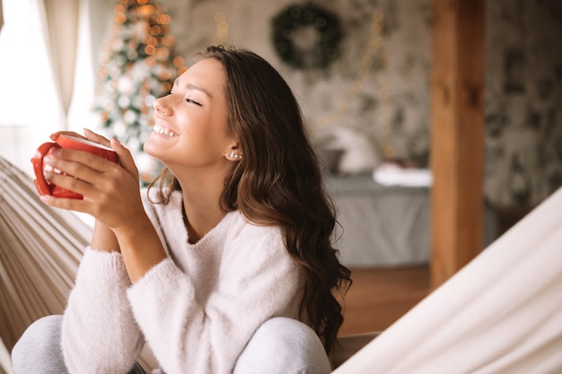 Sorridente ragazza dai capelli scuri vestita con maglione beige e pantaloni tiene una tazza rossa seduta su un'amaca in un'accogliente stanza decorata con un albero di Capodanno. .