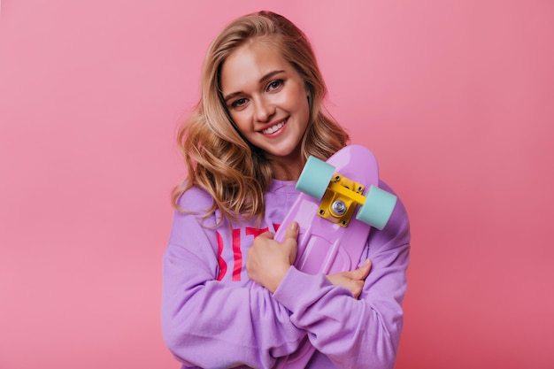 Sorridente ragazza di buon umore con capelli biondi in piedi su sfondo roseo Modello femminile ispirato con skateboard che esprime emozioni felici