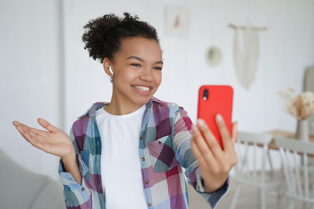 Sorridente ragazza di razza mista blogger in auricolare che chatta tramite videochiamata al telefono conduce aria dal vivo a casa