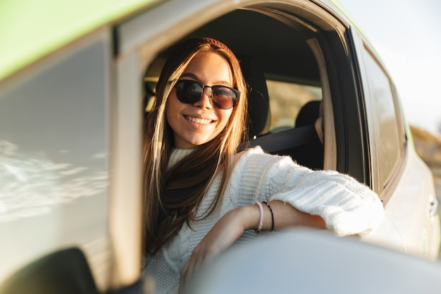 Sorridente ragazza giovane alla guida di un'auto durante il tramonto, guardando la finestra