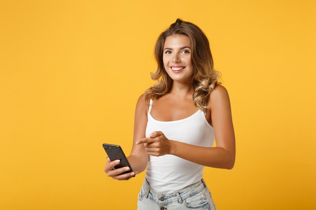 Sorridente ragazza giovane donna in abiti casual leggeri in posa isolata su sfondo giallo arancio. Concetto di stile di vita delle persone. Mock up spazio di copia. Puntare il dito indice sul telefono cellulare, digitare un messaggio sms.