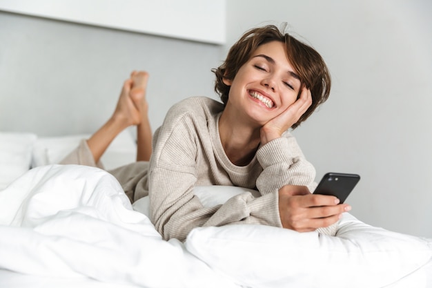 Sorridente ragazza giovane rilassante a letto la mattina, utilizzando il telefono cellulare