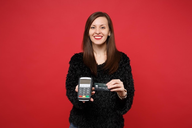 Sorridente ragazza in maglione di pelliccia nera che tiene il terminale di pagamento bancario moderno wireless per elaborare e acquisire pagamenti con carta di credito isolati su sfondo rosso. Concetto di stile di vita di emozioni sincere della gente.