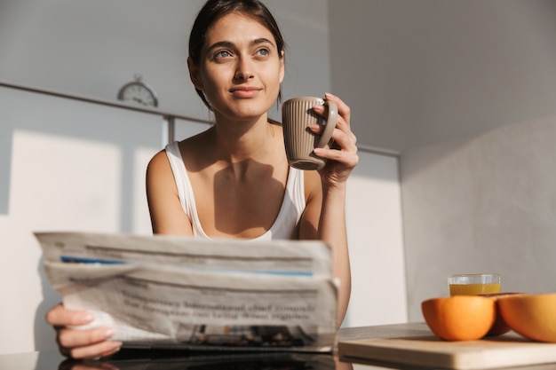 Sorridente ragazza in piedi in cucina al mattino, leggendo il giornale, bevendo il tè