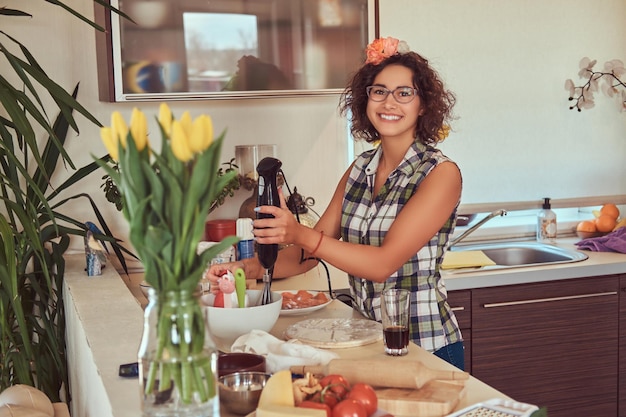 Sorridente ragazza ispanica riccia affascinante che cucina nella sua cucina. Mescola gli ingredienti con una frusta a mano.