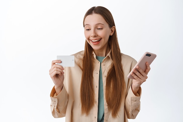 Sorridente ragazza millenaria che tiene in mano uno smartphone, sembra soddisfatta della carta di credito, paga in internet, fa ordini online, fa acquisti con l'applicazione sul telefono, in piedi sul muro bianco