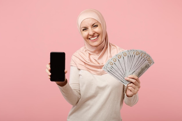 Sorridente ragazza musulmana araba in abiti leggeri hijab in posa isolata su sfondo rosa. Concetto di stile di vita religioso della gente. Mock up spazio di copia. Tenere il cellulare con una ventola vuota dello schermo di denaro contante.