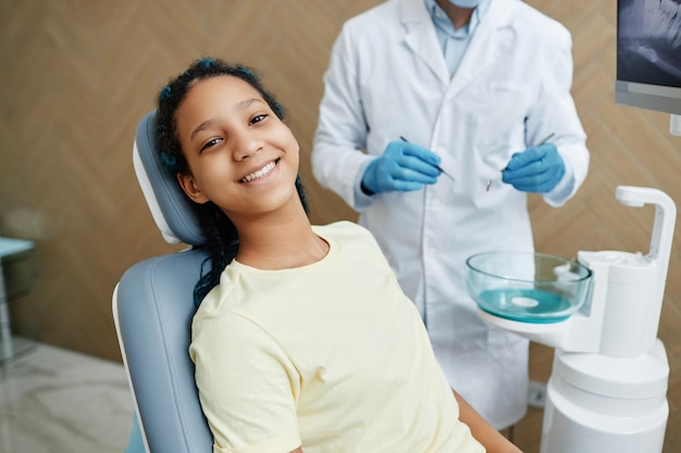 Sorridente ragazza nera seduta nella poltrona del dentista e che guarda l'obbiettivo durante il controllo