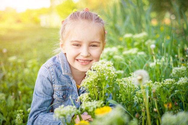 Sorridente ragazza preteen attraente seduta vicino a erba e fiori in giacca di jeans in estate