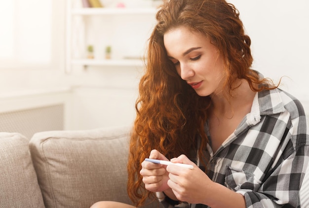 Sorridente ragazza rossa che controlla il suo recente test di gravidanza, seduta sul divano beige a casa, copia spazio