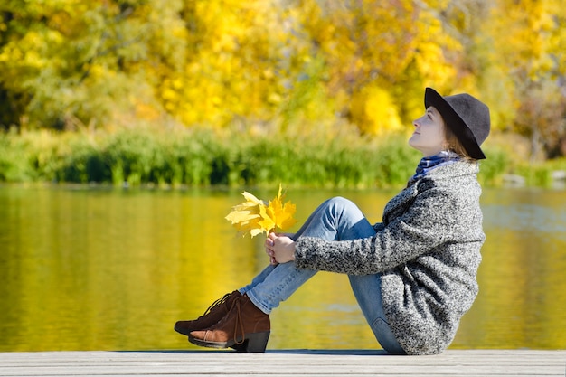 Sorridente ragazza seduta sul molo con foglie d'autunno in mano. Giorno soleggiato