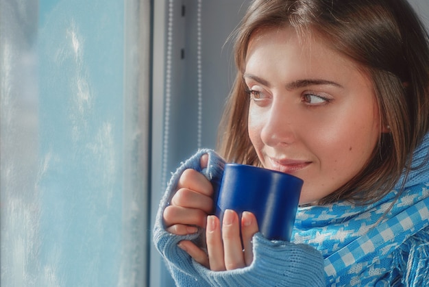 sorridente ragazza sognante in maglione con la tazza vicino alla finestra congelata in inverno