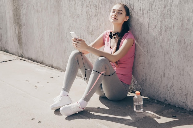 Sorridente ragazza stanca seduta per terra all'aperto che riposa dopo un duro allenamento e acqua potabile