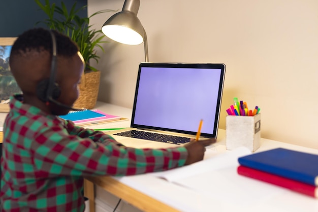 Sorridente ragazzo afroamericano che impara, usando le cuffie e il computer portatile con lo spazio della copia a casa