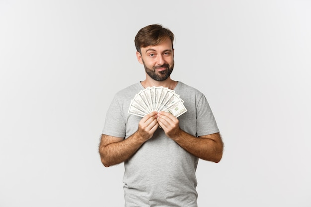 Sorridente ragazzo astuto con la barba, mostrando i soldi, in piedi su sfondo bianco in maglietta grigia