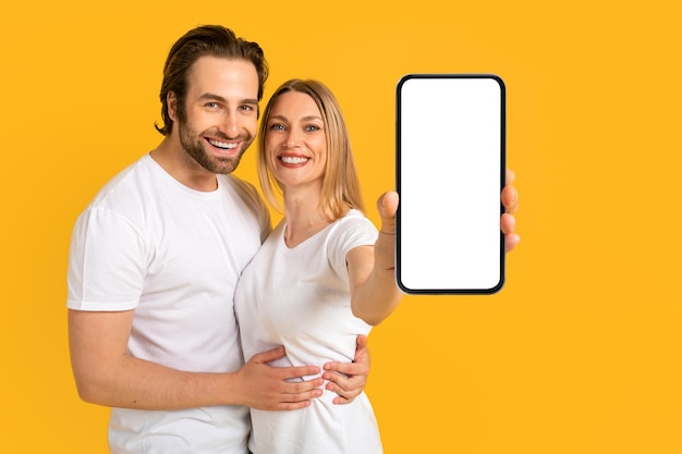 Sorridente ragazzo e signora europei millenari in magliette bianche che abbracciano mostra smartphone con schermo vuoto
