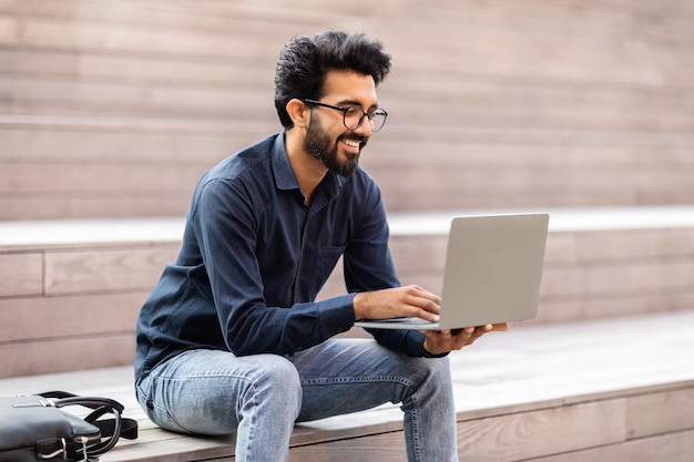 Sorridente ragazzo orientale seduto sulle scale all'aperto utilizzando il computer portatile