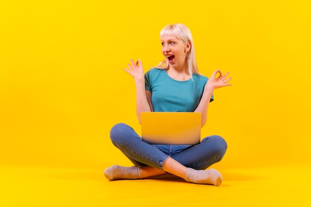 Sorridente seduta con un computer portatile che fa ok bionda ragazza caucasica in studio su sfondo giallo