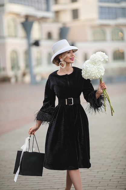 Sorridente signora abbastanza elegante in cappello bianco e vestito nero che cammina per strada. Concetto di strada della moda
