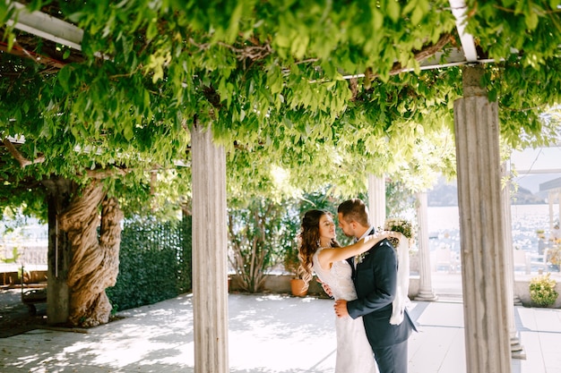 Sorridente sposa abbraccia lo sposo in una giornata di sole in un verde giardino con colonne