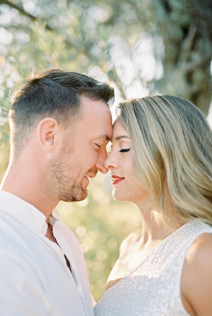 Sorridente sposa e sposo che toccano il naso mentre stanno vicino a un albero verde nel giardino in primo piano