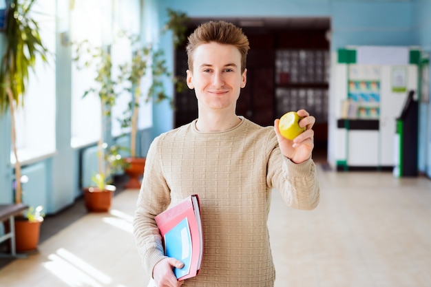 Sorridente studente di college che mangia una mela, in piedi nel corridoio dell'università