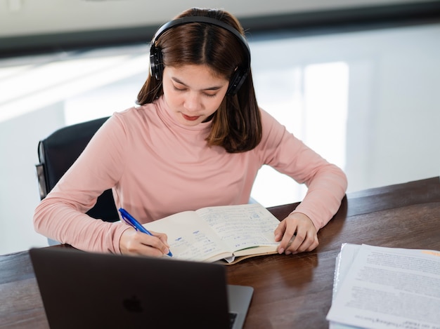 Sorridente studentessa asiatica indossa cuffie wireless scrivere sul notebook per studiare la lingua online guardare e ascoltare il docente, webinar tramite videochiamata e-learning a casa, formazione a distanza