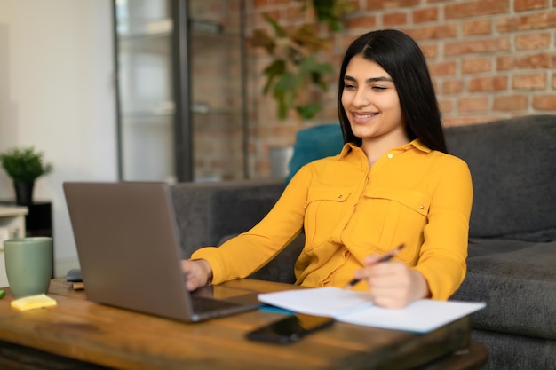 Sorridente studentessa messicana che usa il computer portatile e scrive sul taccuino guardando la lezione studiando online a casa