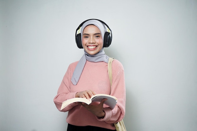 Sorridente studentessa musulmana asiatica in maglione rosa con borsa mentre si ascolta musica e si legge un libro