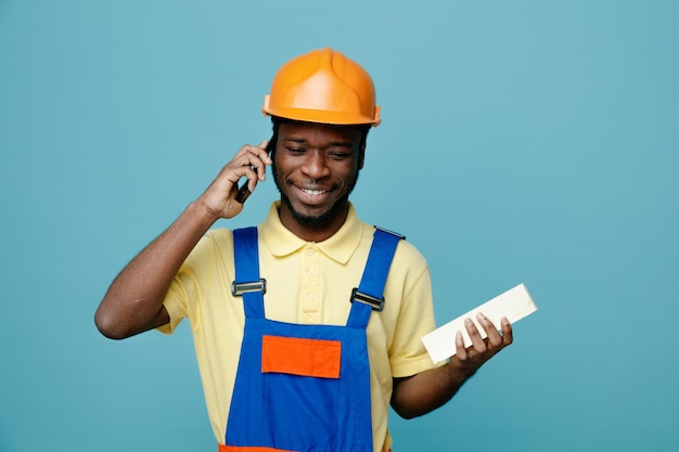 Sorridente tenendo il mattone parla al telefono giovane costruttore afroamericano in uniforme isolato su sfondo blu